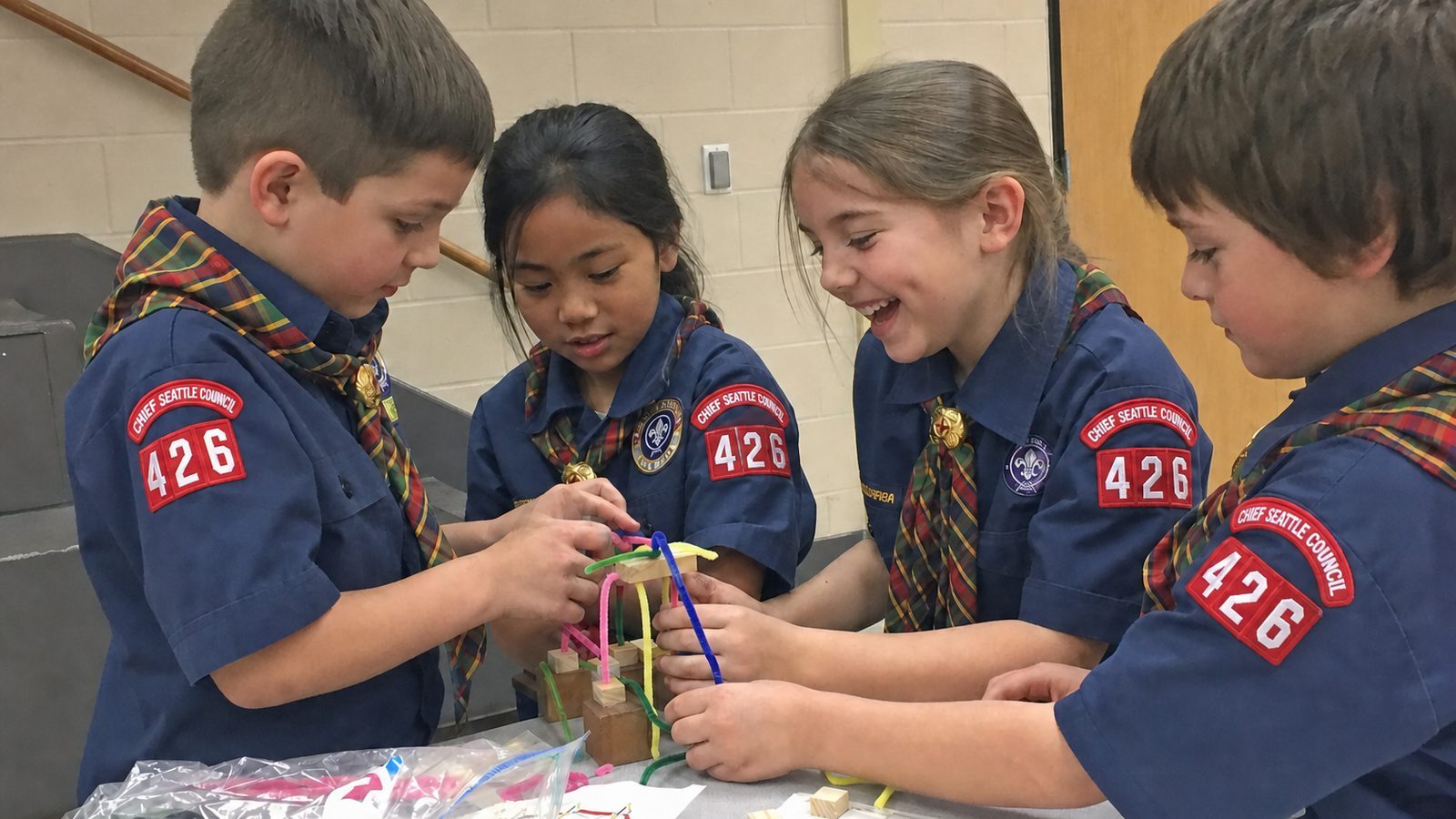 Four Cub Scouts leaning over a table, grinning, building something out of pipe cleaners and wooden blocks at a pack meeting.
